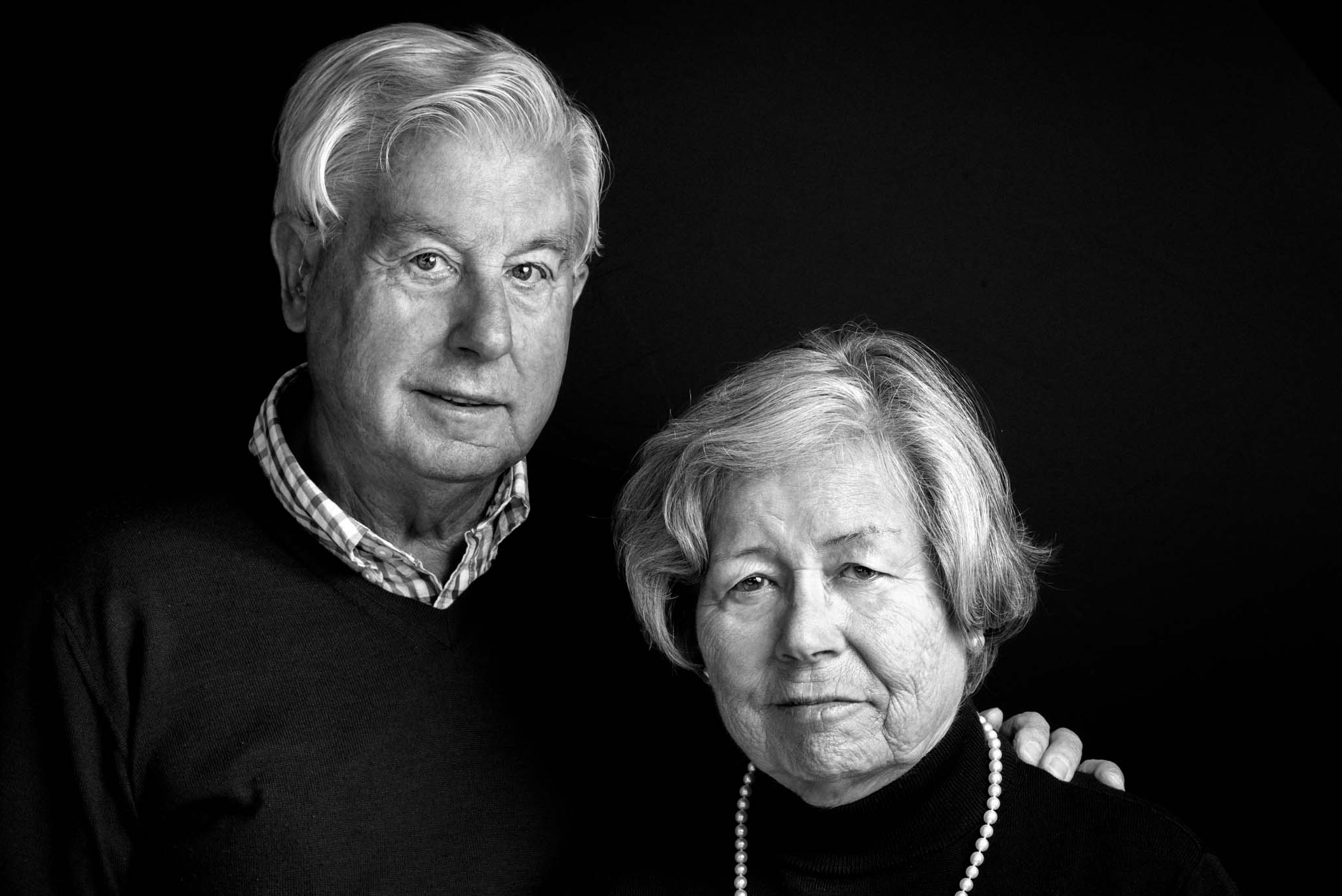 Portrait von einem älteren Ehepaar in schwarzweiß. Beide schauen in die Kamera. Der Mann legt seinen Arm um die Frau.