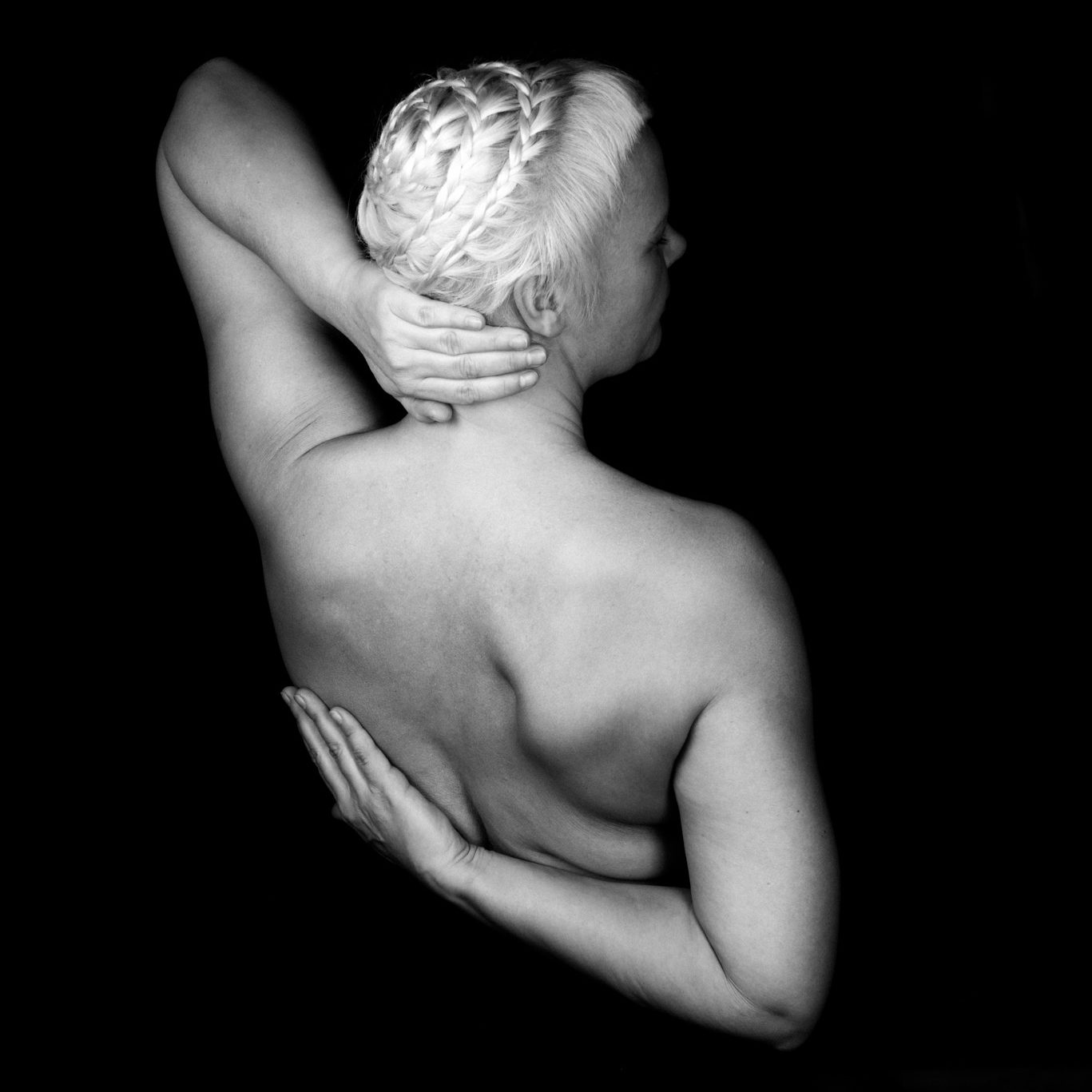 Portraitfoto von einer Frau von Hinten. Wir sehen ihren Rücken und ihre Arme. Ihre Haare sind eigenwillig zu einem runden Zopf geflochten. Ihre linke Hand hält ihren nacken, der rechte arm ist hinter dem Rücken verschränkt.