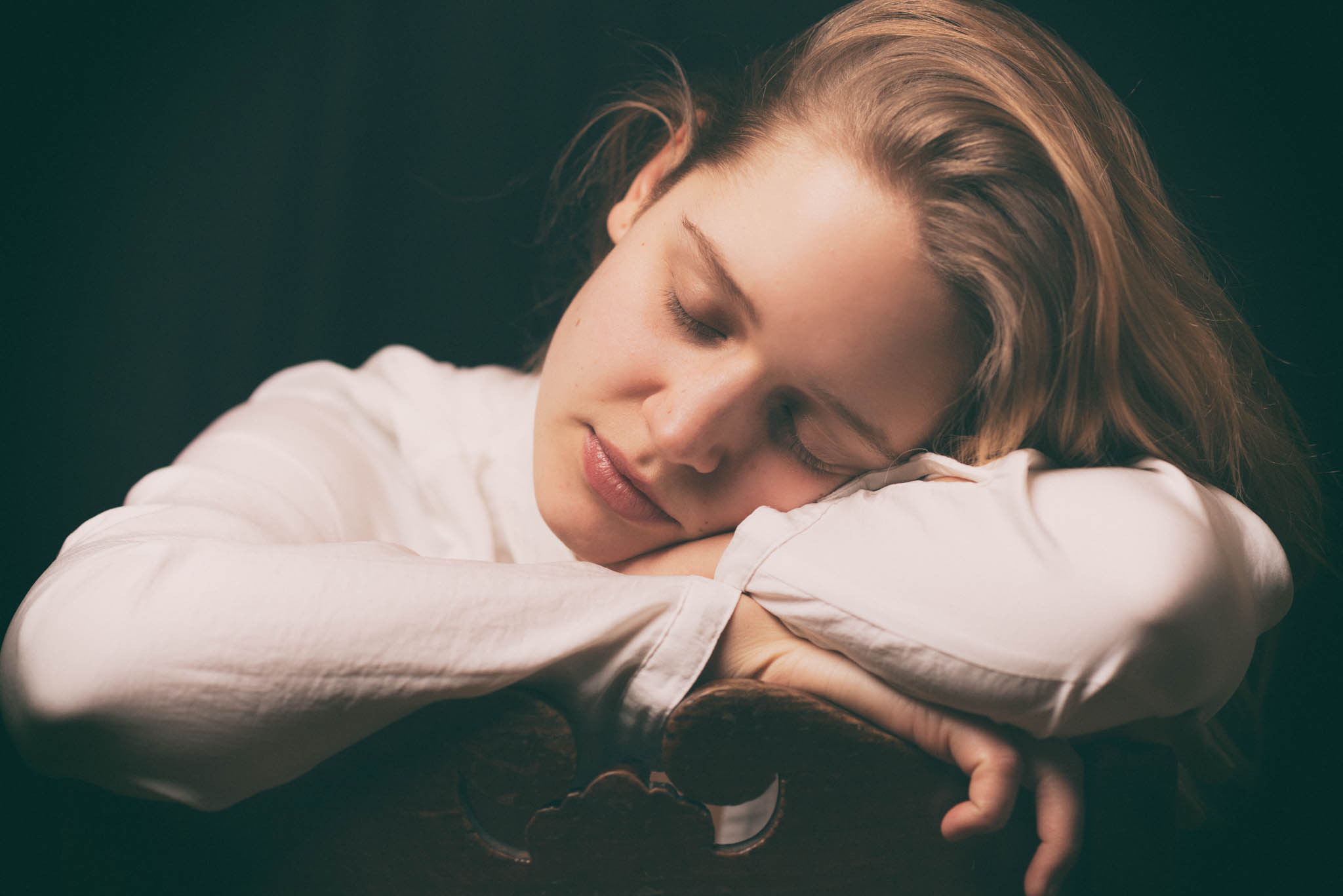 Portraitaufnahme einer schlafenden, jungen Frau. Es ist ihr Kopf zu sehen, den sie auf ihren Armen abgelegt hat, die wiederum auf einer Stuhllehne aufliegen. Sie wirkt sehr friedlich und rein in ihrer Pose und der weißen Bluse.