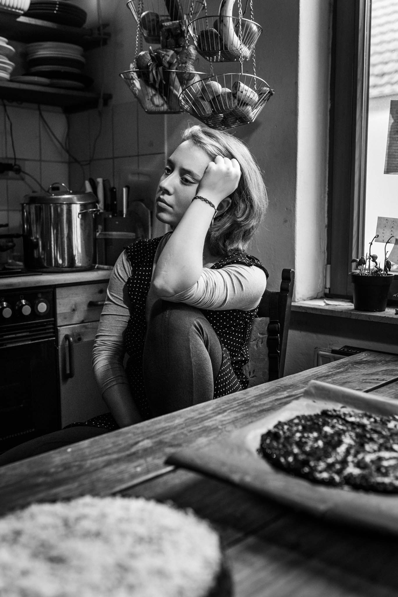 Foto einer jungen Frau in ihrer Küche. Sie sitzt auf einem Stuhl, ein Bein angezogen, den Kopf mit dem Arm auf das Knie gestützt. Ihr Blick ist gedankenversunken. Vor ihr stehen zwei frisch gebackene Kuchen.