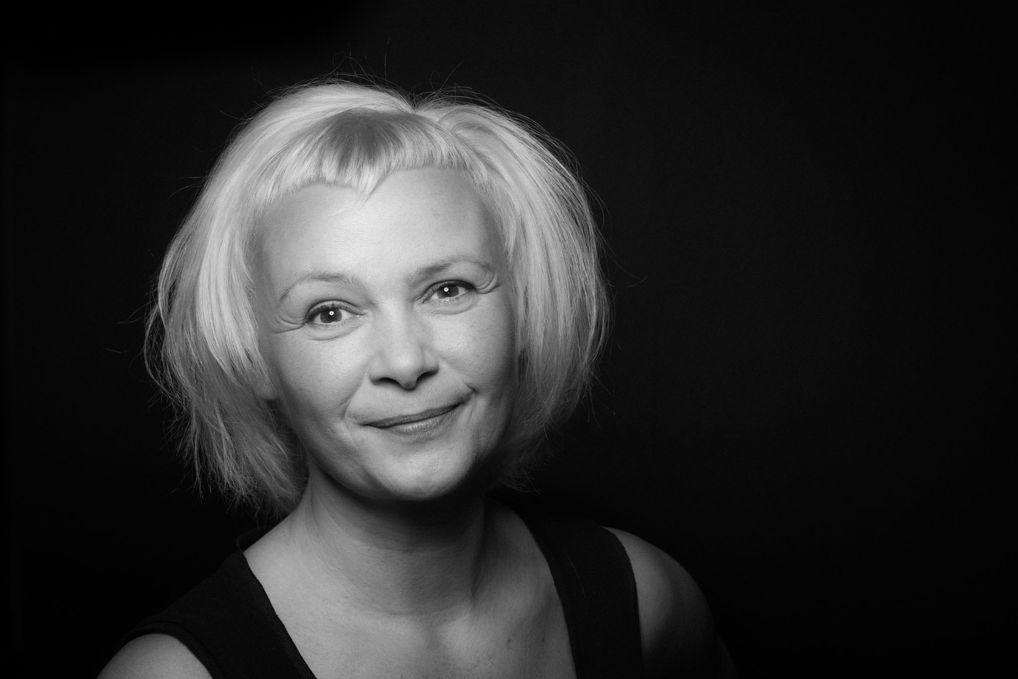 Portraitaufnahme einer Frau mit weißem Bubikopf. In schwarzweiß wirkt die Aufnahme sehr kontrastreich. Sie schaut freundlich, mit einem leichten Lächeln in die Kamera.